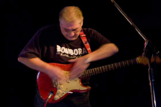 A man wearing a black tshirt reading ‘BOMBORA’ and playing a red electric guitar. His arms are blurred due to motion.